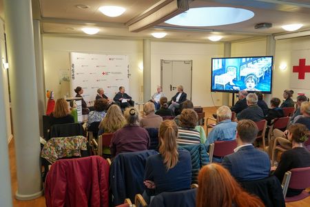 251007_Vernissage-KEINORT Mehrere Dutzend Menschen schauen sich eine Gesprächsrunde von 5 Menschen an.