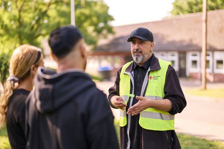 Symbolbild Unterkünfte für Geflüchtete Drei Personen unterhalten sich. Eine von ihnen trägt eine gelbe Warnweste mit DRK-Logo.