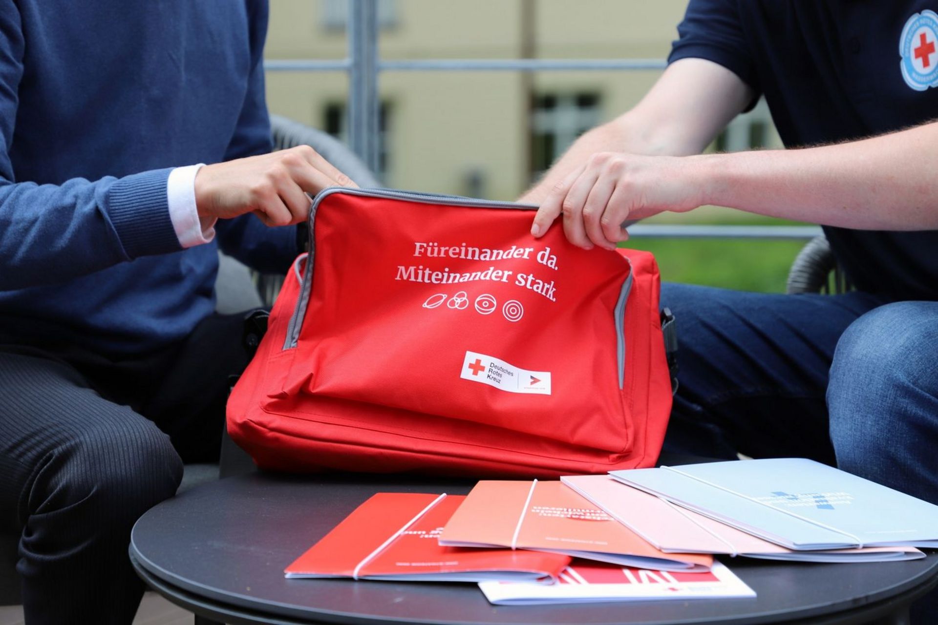 Zwei Mitarbeitende sitzen nebeneinander und greifen in eine DRK-Tasche rein